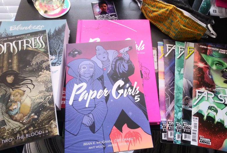 the covers of Monstress, Paper Girls and several issues of Far Sector laid out on a desk. two face masks are at the top right.