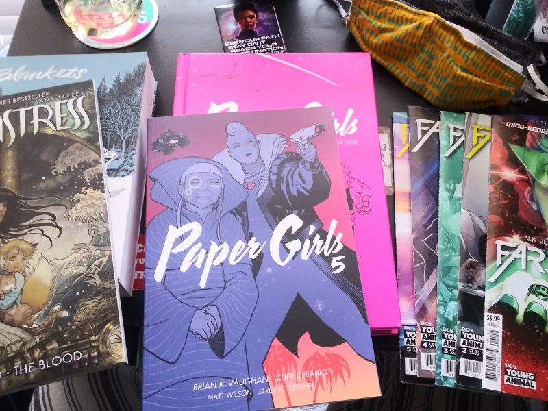 the covers of Monstress, Paper Girls and several issues of Far Sector laid out on a desk. two face masks are at the top right.