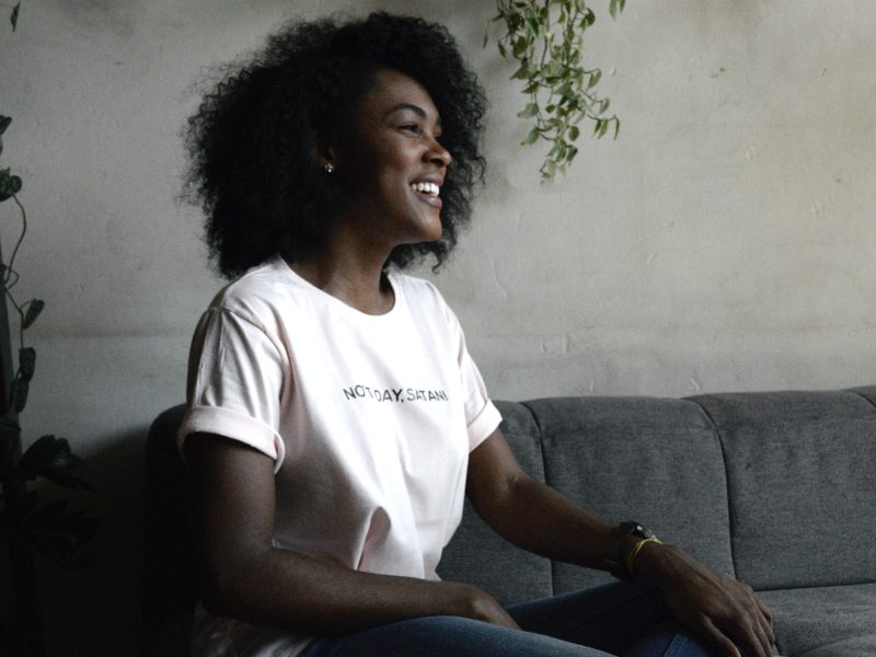 a black woman sits on a grey couch with a smile. her shirt says Not Today, Satan.
