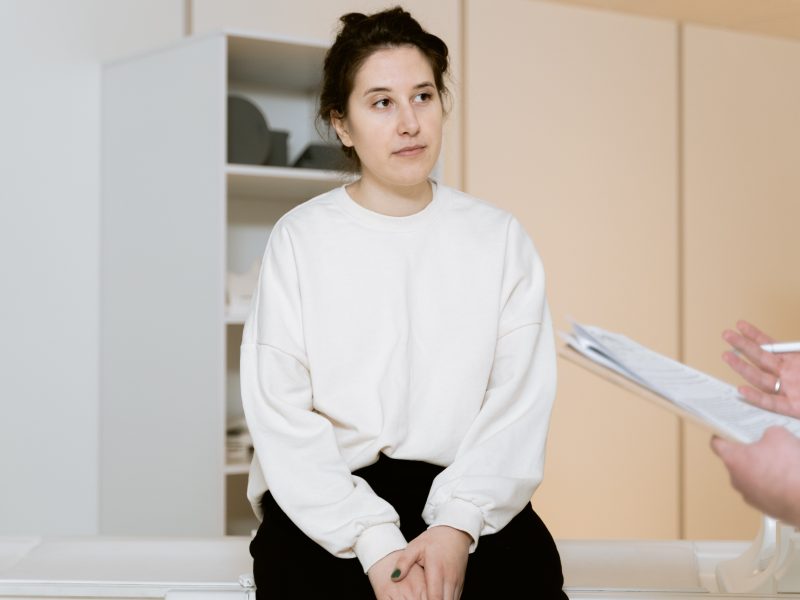 Person in a white sweater and black pants sits on an exam table. A clipboard is being held by someone off screen.