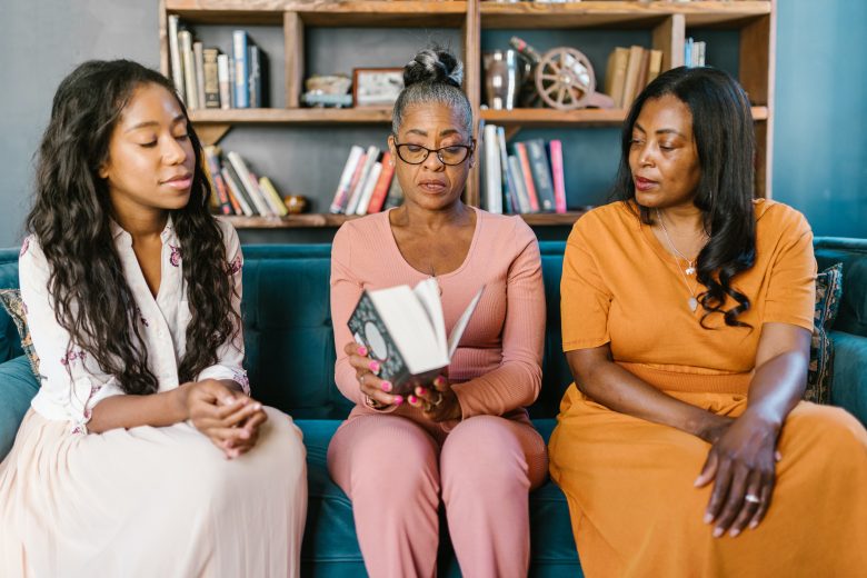 three generations of black women sit on a couch in front of a bookcase. the youngest is on the left, watching a grey-haired woman as she reads. the last woman on the right looks intently.