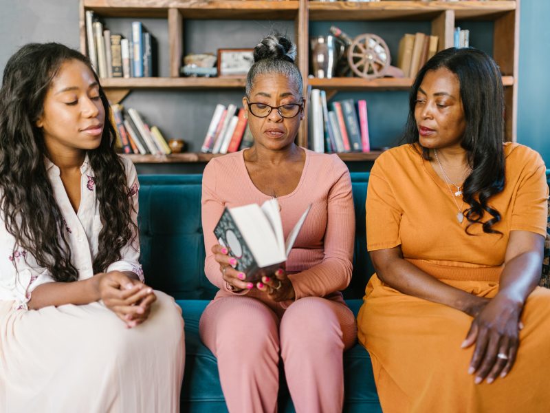 three generations of black women sit on a couch in front of a bookcase. the youngest is on the left, watching a grey-haired woman as she reads. the last woman on the right looks intently.