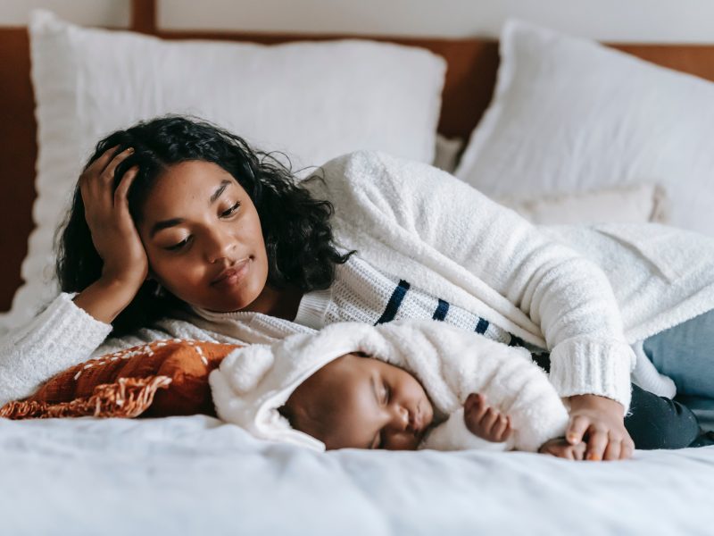a black woman props herself up on one arm while laying on a bed, with a baby sleeping under her left arm. she is looking down at the child lovlingly.