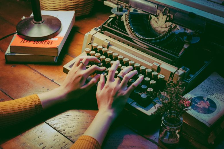 two hands rest on top of a vintage typewriter. two books hold up an illuminated light on the left. there's also another stack of books on the right.