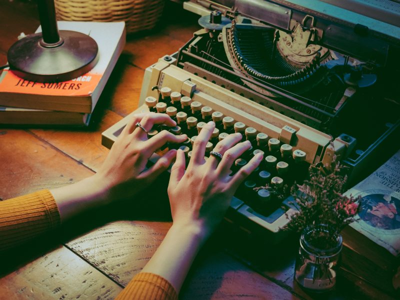 two hands rest on top of a vintage typewriter. two books hold up an illuminated light on the left. there's also another stack of books on the right.