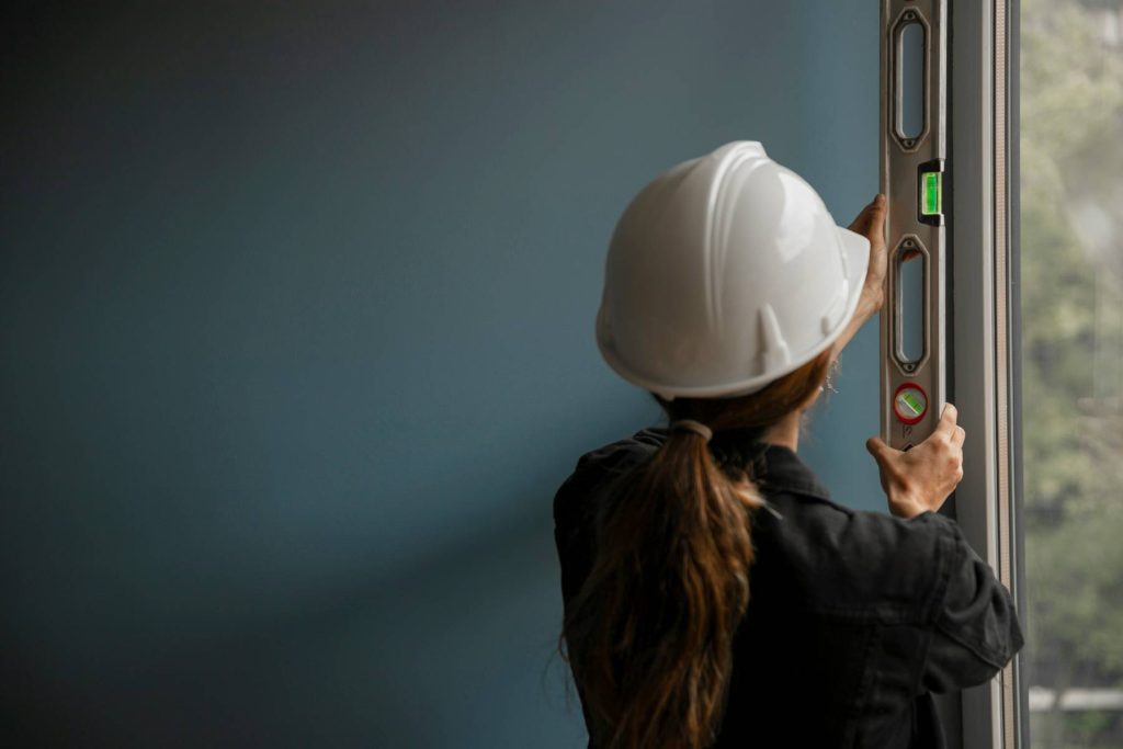 a-woman-in-a-hard-hat-is-looking-out-a-window holding a level